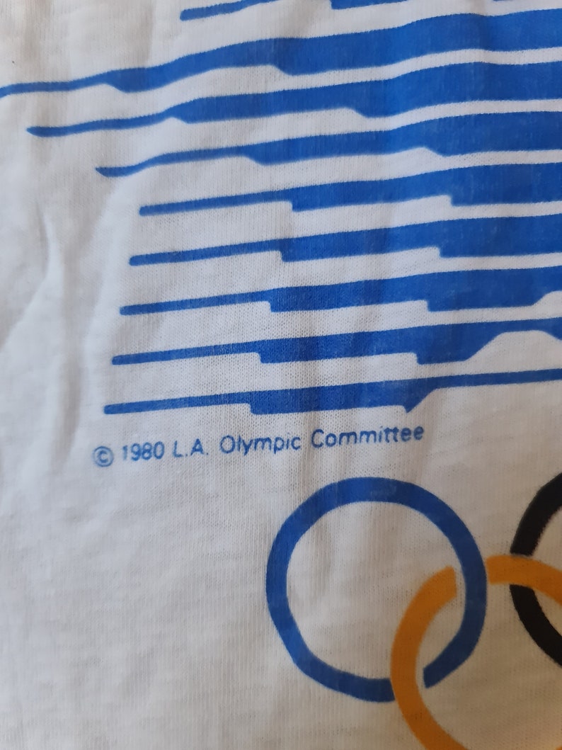 May include: White t-shirt with a blue and white striped design. The shirt has the text "&copy; 1980 L.A. Olympic Committee" and the Olympic rings in blue, yellow, black, and green.