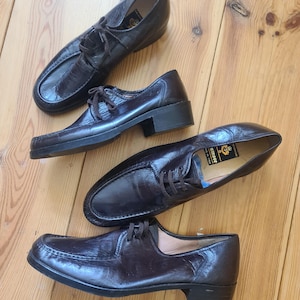 以下が含まれることがあります： 薄茶色の木製の表面に、紐付きの濃い茶色の革製ドレスシューズが4足並んでいます。靴は、低いヒールとテクスチャード加工が施されたクラシックなデザインです。靴は良い状態です。