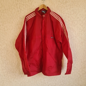 Puede incluir: Una chaqueta cortavientos roja con rayas blancas en las mangas y un logotipo de Adidas negro en el pecho.