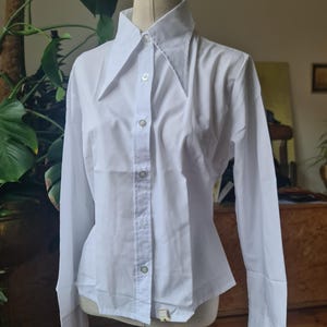 May include: A white long-sleeve button-down shirt with a wide collar. The shirt is displayed on a mannequin. The sleeves have buttoned cuffs. The shirt is made of a smooth, opaque fabric.