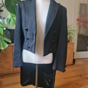 Op de afbeelding: Zwarte smokingjas met fluwelen kraag en staarten, gepresenteerd op een mannequin. De jas heeft een dubbele rij knopen aan de voorkant met zwarte knopen. Het kledingstuk is op maat gemaakt en lijkt van een donkere stof te zijn.