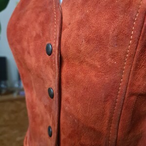 May include: Close-up of a rust-colored suede vest with a V-neck and four black buttons down the front. The vest has visible stitching along the seams and a soft, textured appearance.