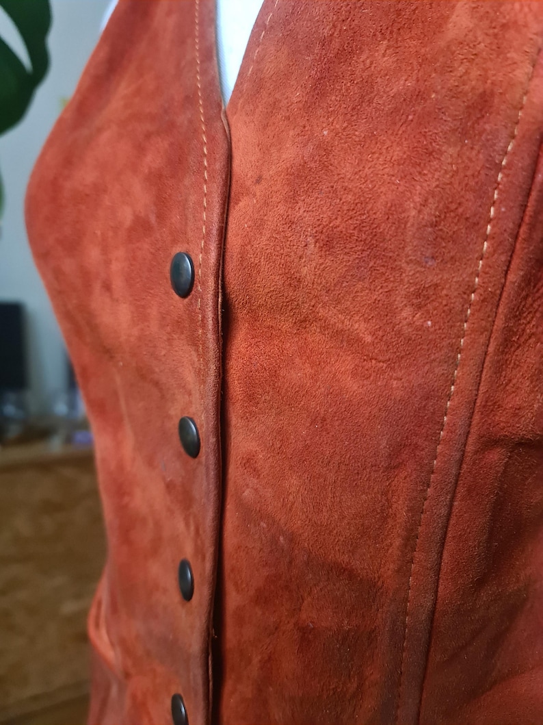 May include: Close-up of a rust-colored suede vest with a V-neck and four black buttons down the front. The vest has visible stitching along the seams and a soft, textured appearance.