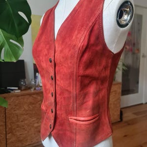 May include: A vintage red suede vest with a V-neck and button closure. The vest features a small pocket on the front and is displayed on a mannequin. The vest's color is a warm, reddish-orange hue.