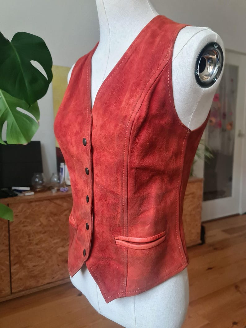 May include: A vintage red suede vest with a V-neck and button closure. The vest features a small pocket on the front and is displayed on a mannequin. The vest's color is a warm, reddish-orange hue.