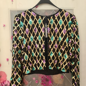 Könnte beinhalten: Eine schwarze Paillettenjacke mit einem Rautenmuster in Gold, Türkis und Lila. Die Jacke hat einen Reißverschluss vorne und lange Ärmel.
