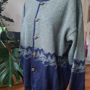 May include: A green and navy blue cardigan. The sweater features a mountain design at the bottom and navy blue collar and cuffs. It is fastened with gold-coloured buttons. The garment is displayed on a mannequin.