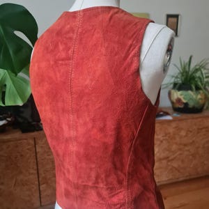 May include: A rust-colored suede vest displayed on a mannequin. The vest has a V-neck and a tailored fit. The back of the vest is visible, showing vertical stitching down the center. The garment is a vintage style.