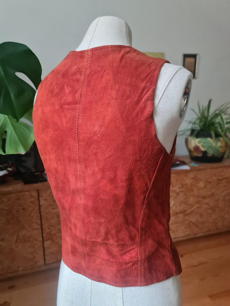 May include: A rust-colored suede vest displayed on a mannequin. The vest has a V-neck and a tailored fit. The back of the vest is visible, showing vertical stitching down the center. The garment is a vintage style.