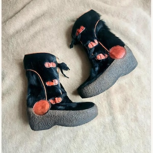 Pode incluir: Um par de botas de cano médio em material preto com textura de pelo, acentuadas com debruns, remendos circulares e fechos de alavanca cor de laranja. As botas têm uma sola de borracha espessa cinzento-escura com um acabamento granulado.