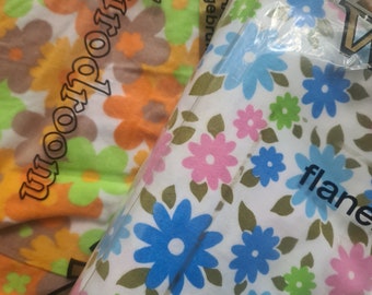 Puede incluir: Dos paquetes de tela con estampados florales. Un paquete tiene un fondo blanco con flores azules, rosas y verdes. El otro paquete tiene un fondo blanco con flores naranjas, amarillas y verdes. El texto "Eurotoom" está impreso en el paquete naranja y amarillo. El texto "flanel" está impreso en el paquete blanco y azul.
