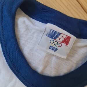 May include: White t-shirt with a blue ribbed collar and a Levi's tag with the Olympic rings and USA logo.
