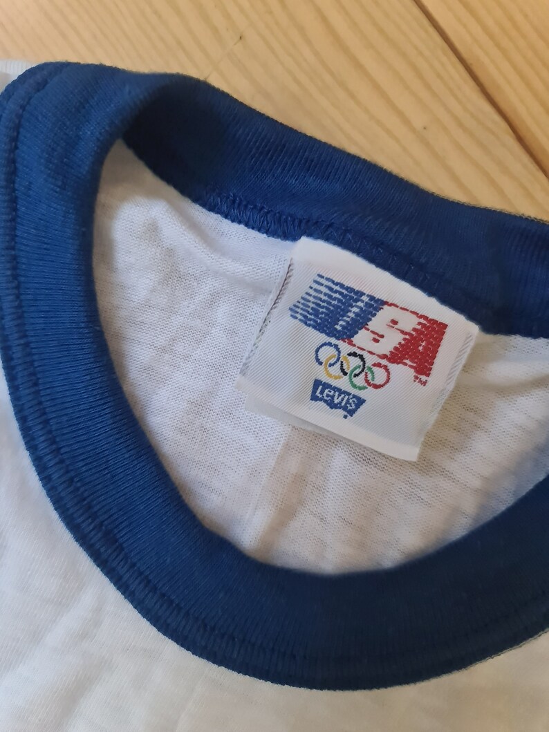 May include: White t-shirt with a blue ribbed collar and a Levi's tag with the Olympic rings and USA logo.