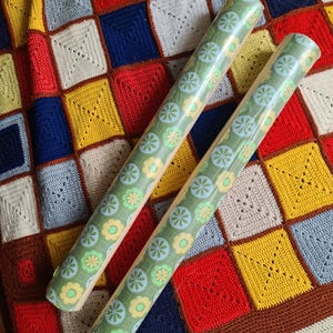Puede incluir: Dos rollos de papel de regalo verde con un estampado floral de flores azul claro y amarillas. Los rollos est&aacute;n sobre una colorida manta de ganchillo patchwork con cuadrados rojos, amarillos, azules y blancos.