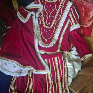 May include: A red velvet costume with gold trim, consisting of a tunic, pants, and cape. The tunic has a high collar and is adorned with gold embroidery and gemstones. The pants have vertical gold stripes.