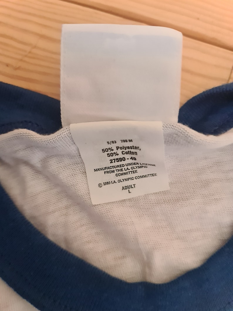 May include: White tag with black text, 5/83 788 M 50% Polyester, 50% Cotton 27590-49 MANUFACTURED UNDER LICENSE FROM THE LA. OLYMPIC COMMITTEE &copy; 1980 LA. OLYMPIC COMMITTEE ADULT L