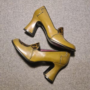 May include: A pair of mustard yellow heeled shoes with a leopard print detail on the back. The shoes have a chunky heel and a pointed toe.