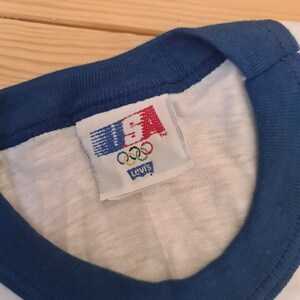 May include: A white t-shirt with a blue ringer neck and a Levi's tag that says "USA" with the Olympic rings and "Levi's" underneath.