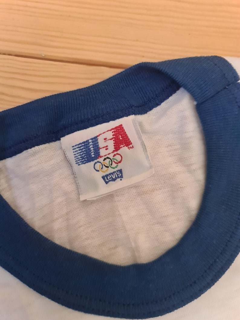 May include: A white t-shirt with a blue ringer neck and a Levi's tag that says "USA" with the Olympic rings and "Levi's" underneath.