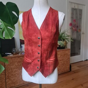 May include: A rust-colored suede vest with a V-neck and a button-down front. The vest has two small pockets and is displayed on a mannequin. The vest is a vintage style and appears to be in good condition.