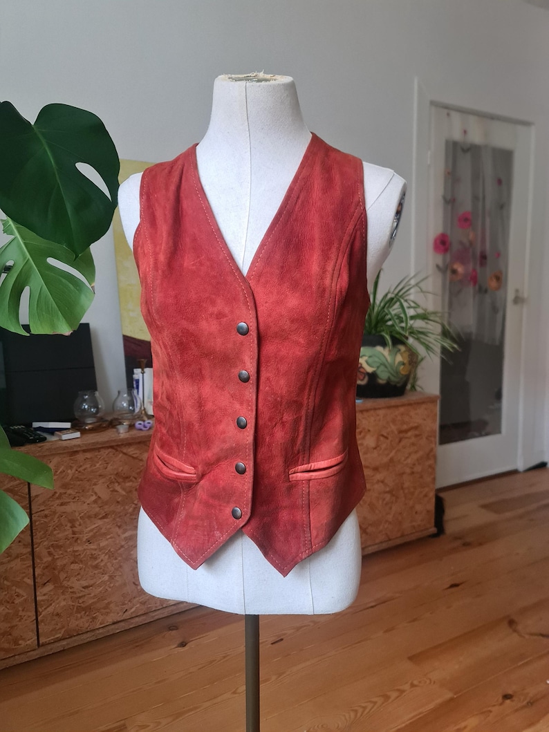 May include: A rust-colored suede vest with a V-neck and a button-down front. The vest has two small pockets and is displayed on a mannequin. The vest is a vintage style and appears to be in good condition.