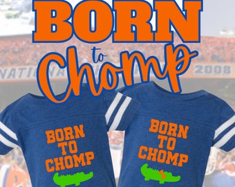 May include: Two blue t-shirts with white stripes on the sleeves. Each shirt has the words "BORN TO CHOMP" in orange, with a green alligator graphic. The phrase "BORN to Chomp" is also written in orange and blue above the shirts.
