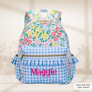 May include: A backpack featuring a white base with a colorful floral pattern of pink, yellow, and blue flowers. The front pocket is light blue and white gingham with a ruffled trim and the name Maggie printed in pink. It has gold-tone zippers.