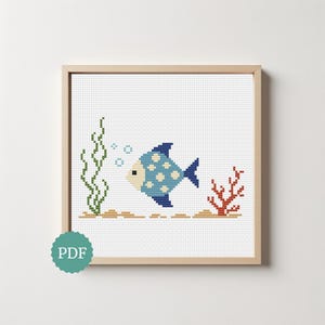 May include: A cross-stitch pattern featuring a blue fish with white polka dots, swimming near green seaweed and red coral on a white grid background. The design includes small blue bubbles and a tan sandy bottom, all framed in a light wood square frame.