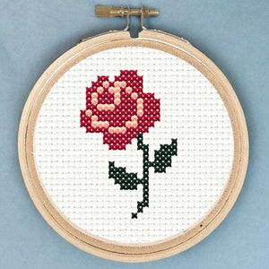 May include: A red and white cross-stitch embroidery of a single rose with green leaves in a wooden embroidery hoop.