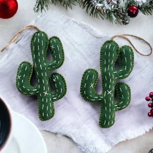 Può includere: Due decorazioni di cactus in feltro verde con cuciture bianche e un anello per appendere. Le decorazioni sono su uno sfondo di lino bianco.