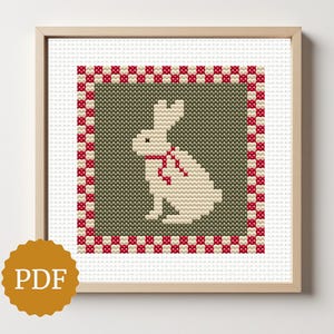 May include: Cross-stitch artwork of a white rabbit with a red bow, set against a green background. The design is framed with a red and white checkered border, presented in a wooden frame. The image includes a PDF icon.