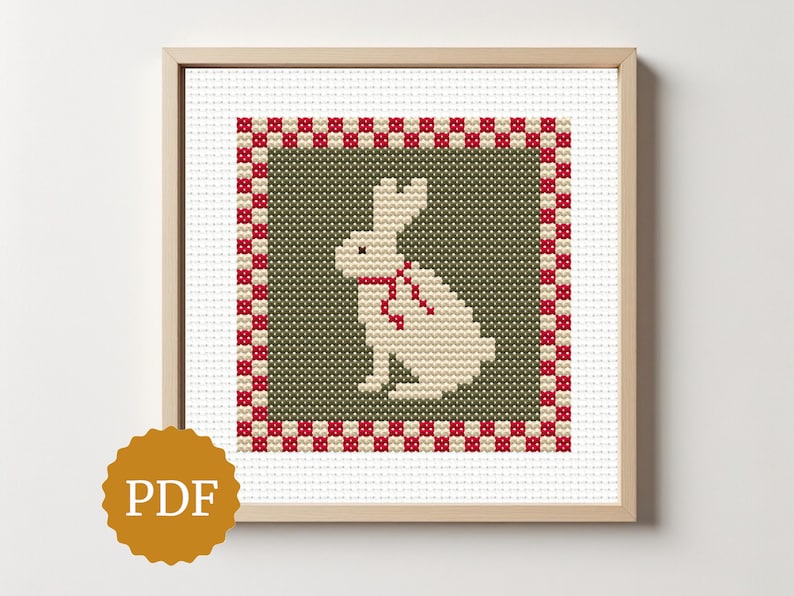 May include: Cross-stitch artwork of a white rabbit with a red bow, set against a green background. The design is framed with a red and white checkered border, presented in a wooden frame. The image includes a PDF icon.