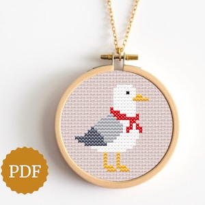 May include: A cross-stitch seagull design in a wooden embroidery hoop pendant. The seagull is white, gray, and black, with a red scarf and yellow feet. The pendant hangs from a gold-colored chain. The image also includes a PDF logo.