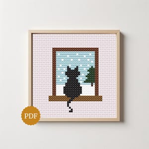 May include: A cross-stitch pattern in a light wooden frame featuring a black cat sitting on a windowsill looking out at a snowy landscape with a green pine tree. The background is a light pink grid, and a gold circular badge with the text PDF is visible.