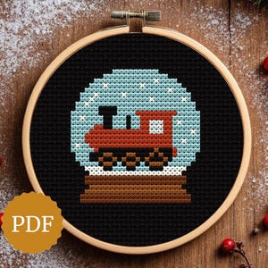 May include: Cross-stitch pattern of a train inside a snow globe, set against a black background. The train is red and black, the snow globe is light blue with white snowflakes. The design is framed in a wooden embroidery hoop. A PDF icon is in the lower left.