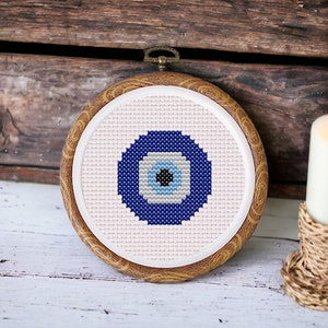 Könnte beinhalten: Ein blaues und weißes böses Auge, das auf eine weiße Leinwand gestickt ist und in einem Holzrahmen eingefasst ist.