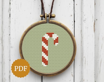 May include: A cross-stitch pendant with a candy cane design. The candy cane is red and white, set against a light green background within a round, bronze-coloured frame. A brown cord is attached for wearing. The image also includes a "PDF" logo.