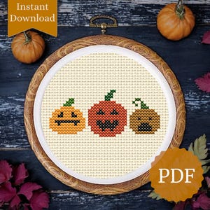 May include: A cross-stitch pattern featuring three smiling pumpkins in orange, red, and brown, each with a green stem. The design is set in a wooden embroidery hoop. The words "Instant Download" and "PDF" are also visible.