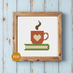 May include: A cross-stitch pattern framed in wood featuring a rust-coloured mug with a cream-coloured heart, a brown steam swirl, and a green book with cream pages. A gold circular PDF icon is visible in the bottom left corner of the frame.