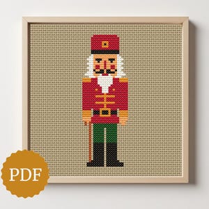 May include: A framed cross-stitch pattern of a nutcracker soldier. The soldier wears a red jacket, green pants, and a black hat. The background is a tan fabric. A gold PDF logo is in the lower left corner.