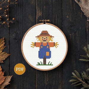 Può includere: Uno schema a punto croce in un telaio da ricamo in legno raffigurante uno spaventapasseri con cappello marrone, camicia arancione e salopette blu. Lo spaventapasseri si trova su un ciuffo d'erba verde. È visibile un badge giallo con testo PDF.