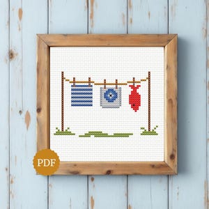Può includere: Un motivo a punto croce incorniciato in legno raffigurante un filo da bucato con tre elementi: un quadrato a righe blu e bianche, un quadrato grigio con un centro blu e una forma di pesce rosso. Il design include pali marroni ed erba verde.