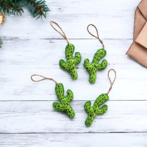 Puede incluir: Cuatro adornos de cactus de fieltro verde con colgadores de cuerda marrón. Los adornos están sobre un fondo de madera blanca.