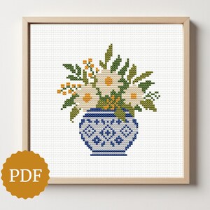 Puede incluir: Patrón de punto de cruz de un arreglo floral en un jarrón azul y blanco. Las flores son blancas con centros amarillos, y las hojas son verdes. El jarrón tiene un diseño geométrico. La obra de arte está enmarcada en un marco de madera clara.