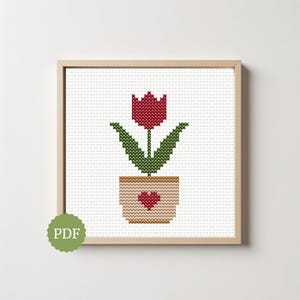 May include: Cross-stitch pattern of a red tulip in a tan pot with a heart, framed in a light wood frame. The tulip has green leaves and is set against a white background. A green PDF logo is in the lower left corner.