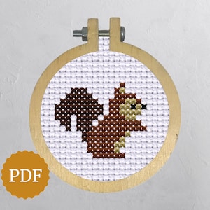 May include: A cross-stitch embroidery of a squirrel in a wooden hoop. The squirrel is brown with a fluffy tail and is stitched on white fabric. The hoop is round and has a metal screw at the top. A gold PDF sticker is in the lower left corner.