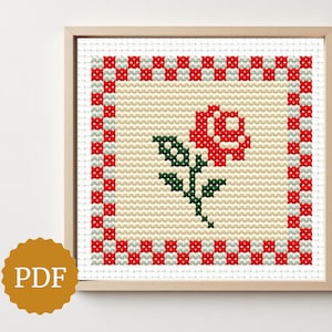 Può includere: Opera d'arte a punto croce di una rosa rossa con foglie e stelo verdi, incorniciata in una cornice di legno. La rosa è centrata su uno sfondo color crema, con un bordo a scacchi rosso e bianco. Un logo PDF è in basso a sinistra.