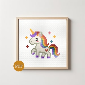 May include: A cross-stitch pattern featuring a white unicorn with a rainbow-colored mane and tail, a yellow horn, and purple hooves. The design includes small colorful stars and a red heart on the unicorn's flank, all framed in a light wood frame.