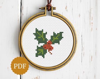 May include: A cross-stitch design of holly leaves and berries, displayed in a round, gold-colored embroidery hoop. The leaves are green, and the berries are red. The hoop is suspended by a brown leather cord. A PDF icon is in the lower left corner.