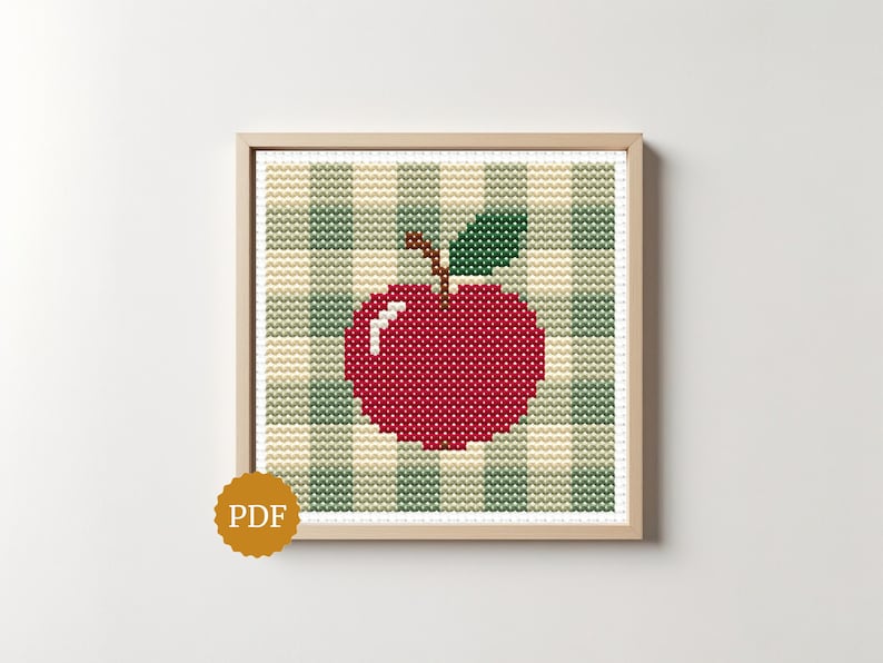 May include: A cross-stitch pattern featuring a bright red apple with a single green leaf and a brown stem, centered on a light beige and sage green gingham checkered background, framed in a light wood square frame with a gold PDF icon.